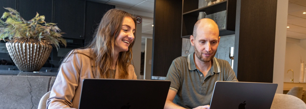 Twee lachende collega's, een man en een vrouw, werken prettig samen achter een laptop in een modern kantoor.